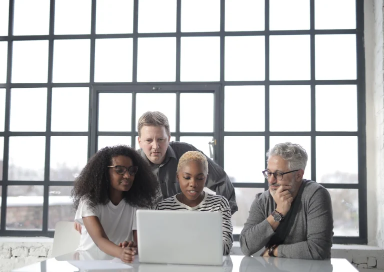 Four colleagues reviewing a laptop together beside large office windows